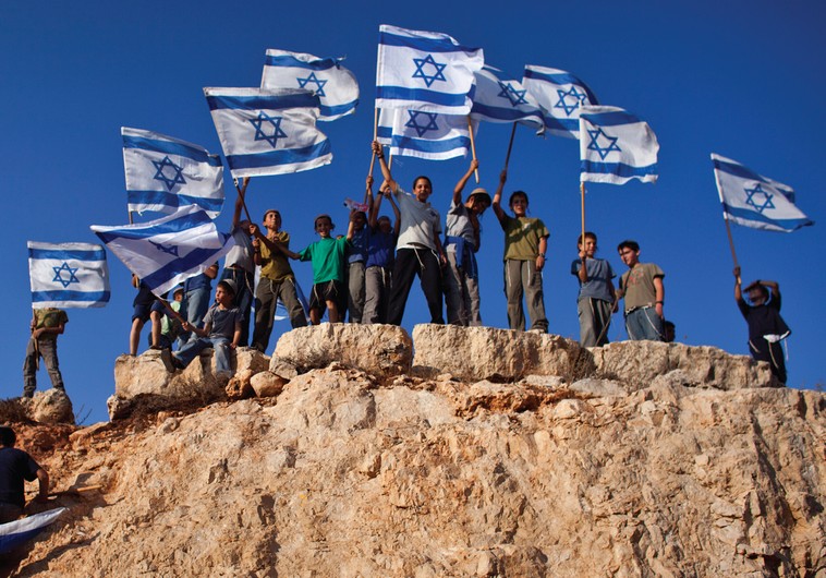 Jewish Youths in Israel wave flags and stand atop a hill. The author recalls his own young days in Zionist youth groups